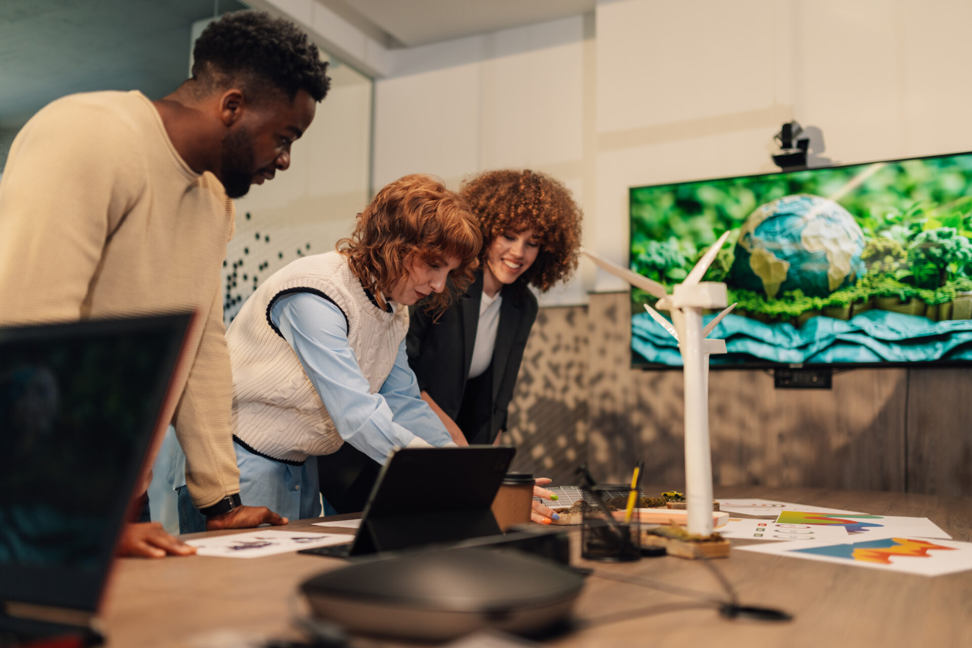Multicultural colleagues standing at eco friendly modern workspace with windmill and solar panels models on table and working with paperwork. Environmentalists discussing sustainability and ecology.