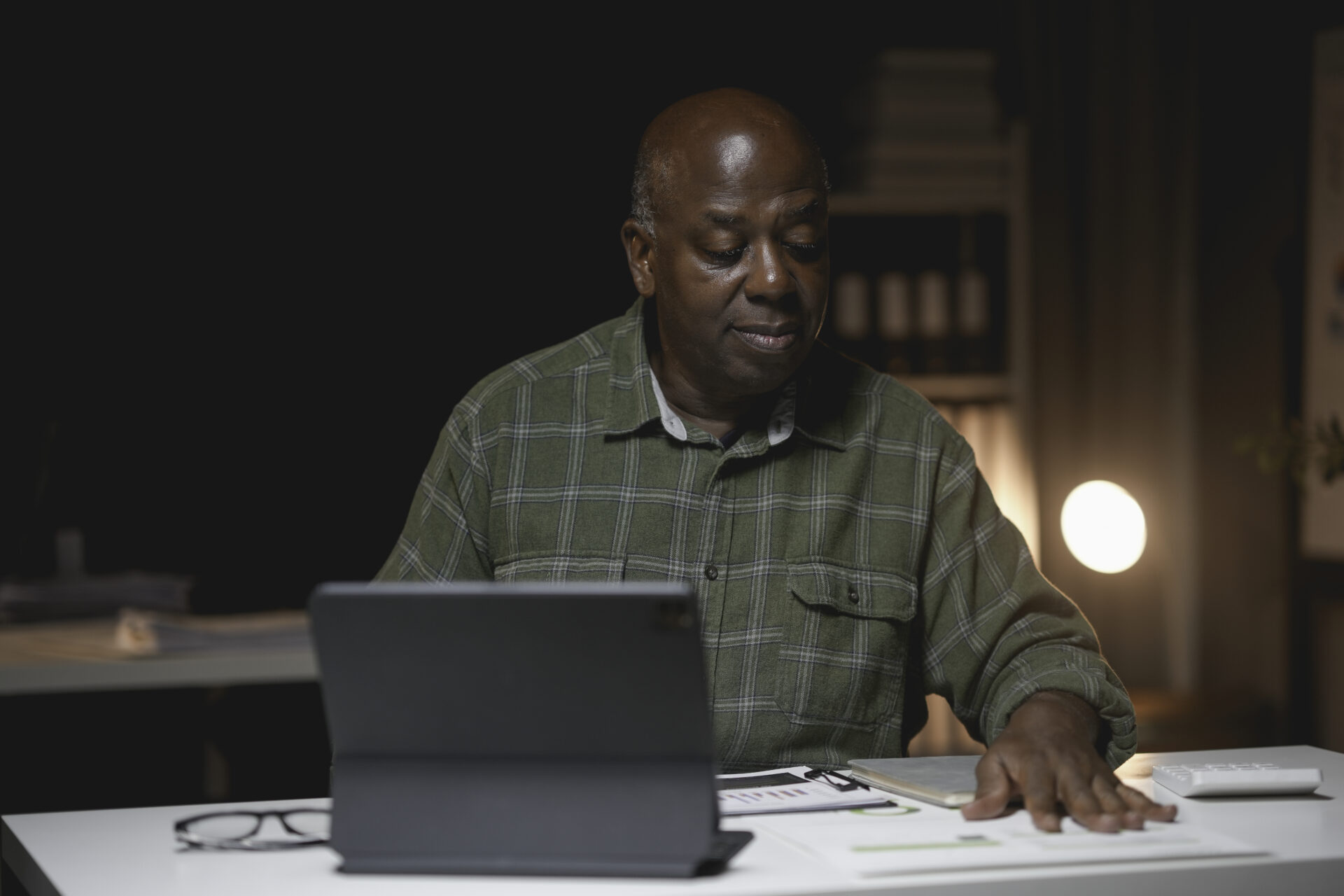 Mature man focuses intently on a digital tablet while working late at his office desk. The warm lighting and organized workspace create a productive atmosphere