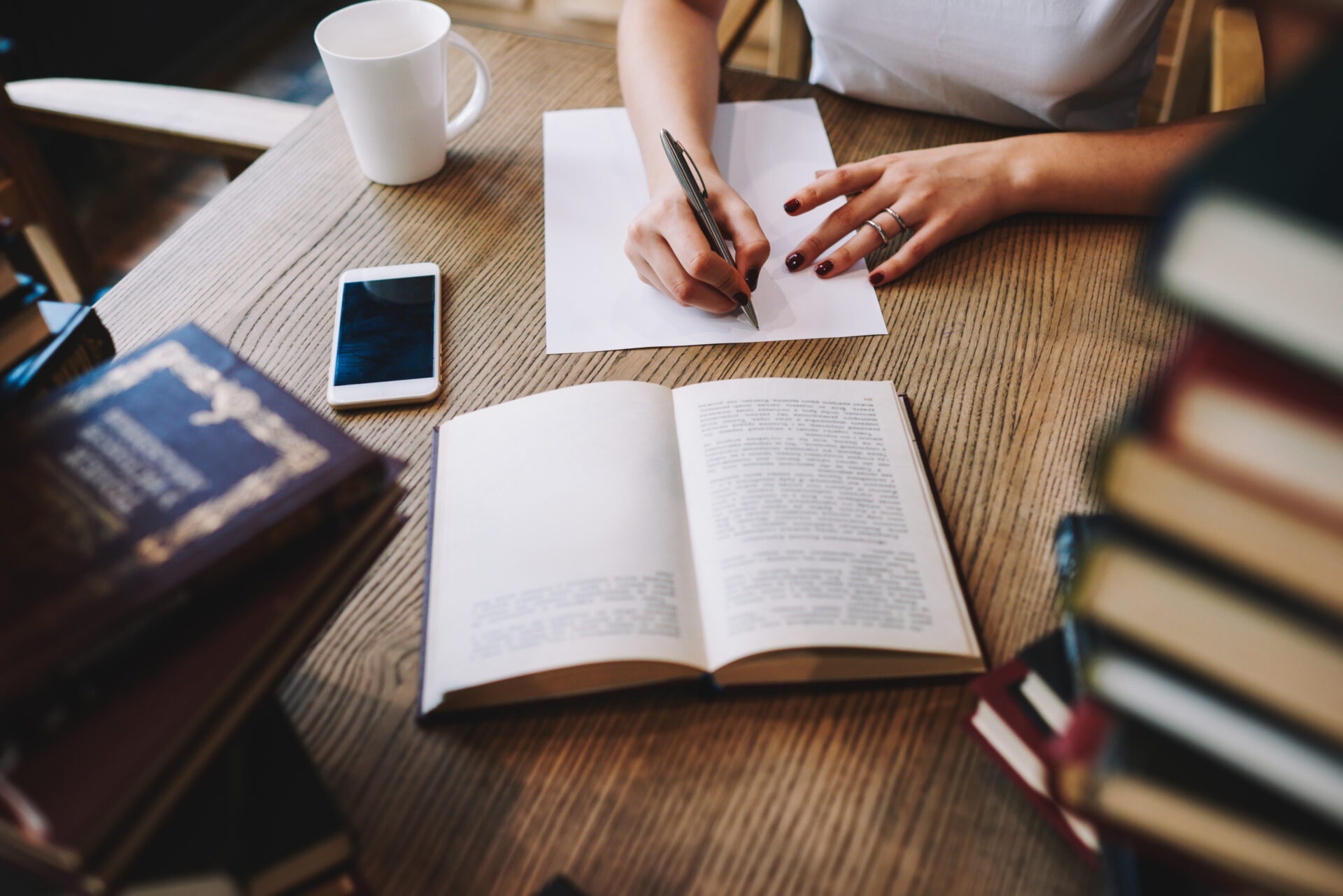 From above of crop faceless female sitting at table with empty cup and smartphone while reading book and writing notes on paper