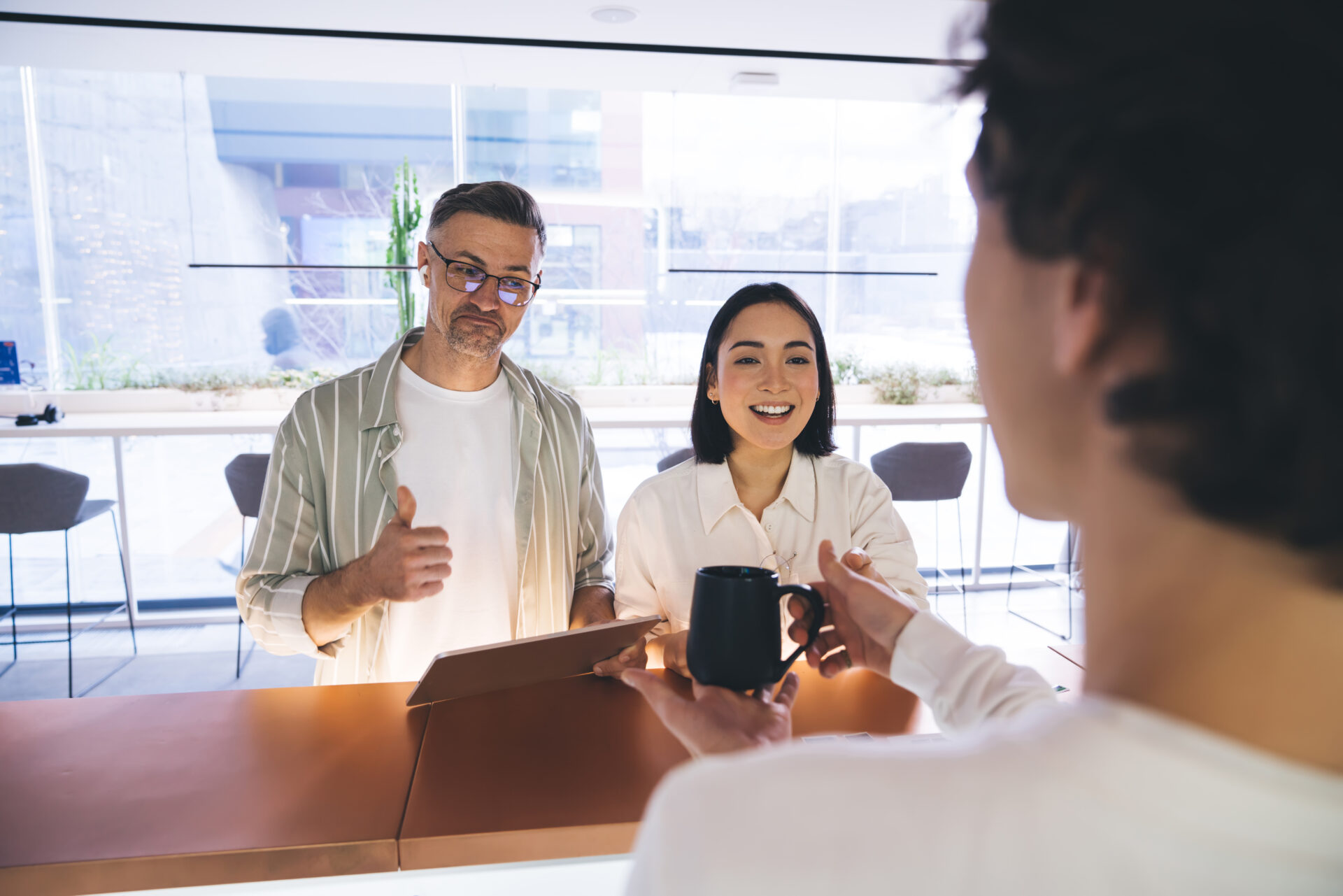 Positive Asian lady in casual clothes and satisfied husband with tablet standing in modern hotel while administrator suggesting cup of coffee